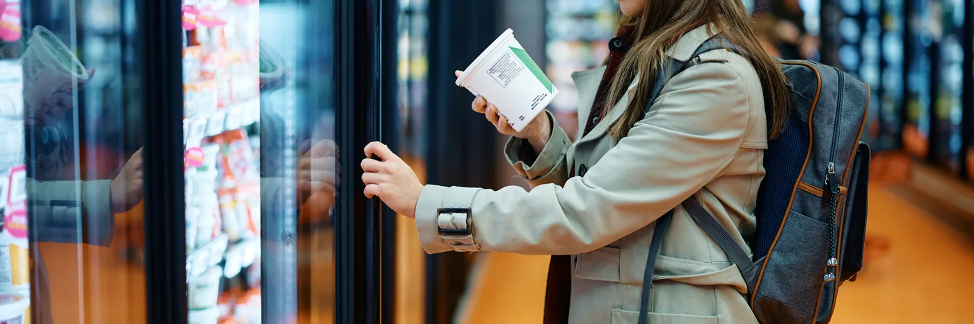 A shopper standing at a supermarket cooler, holding a carton and reading the label on the back.